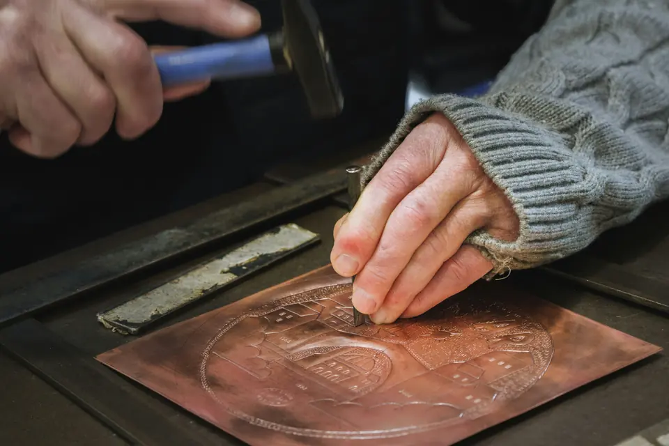 Hands engraving a copper plate with a hammer and a small chisel.