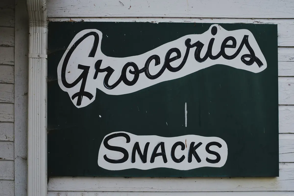 A hand-painted sign on a white weatherboard wall beside a downpipe, with “Groceries” in black script and “Snacks” in capitals on a dark green board.