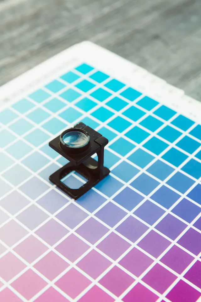 A black printer's loupe standing on a printed chart of cyan to magenta squares.
