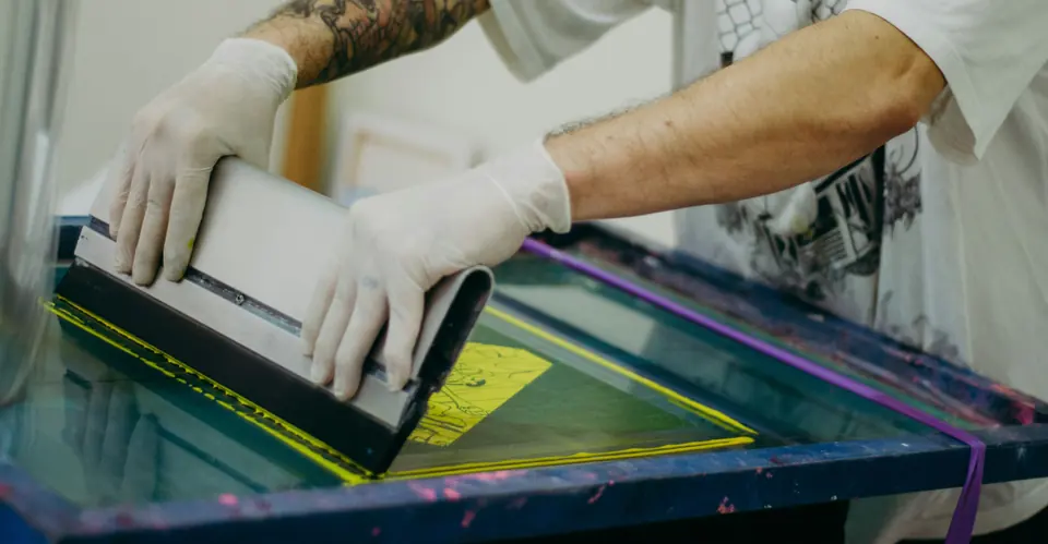 Gloved hands pulling a squeegee across a screen printing frame.