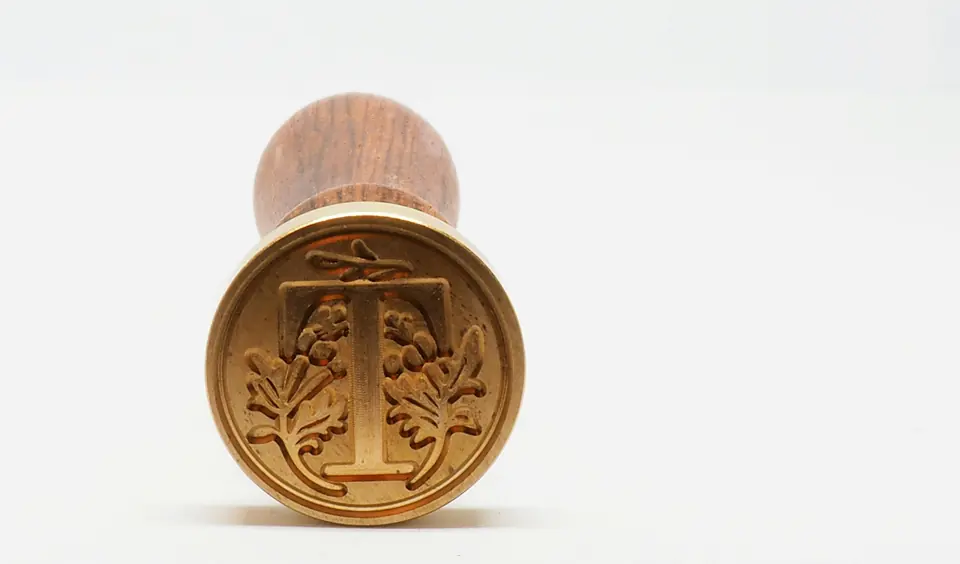 A brass seal stamp with a wooden handle, its face cut with a letter T and leaves.